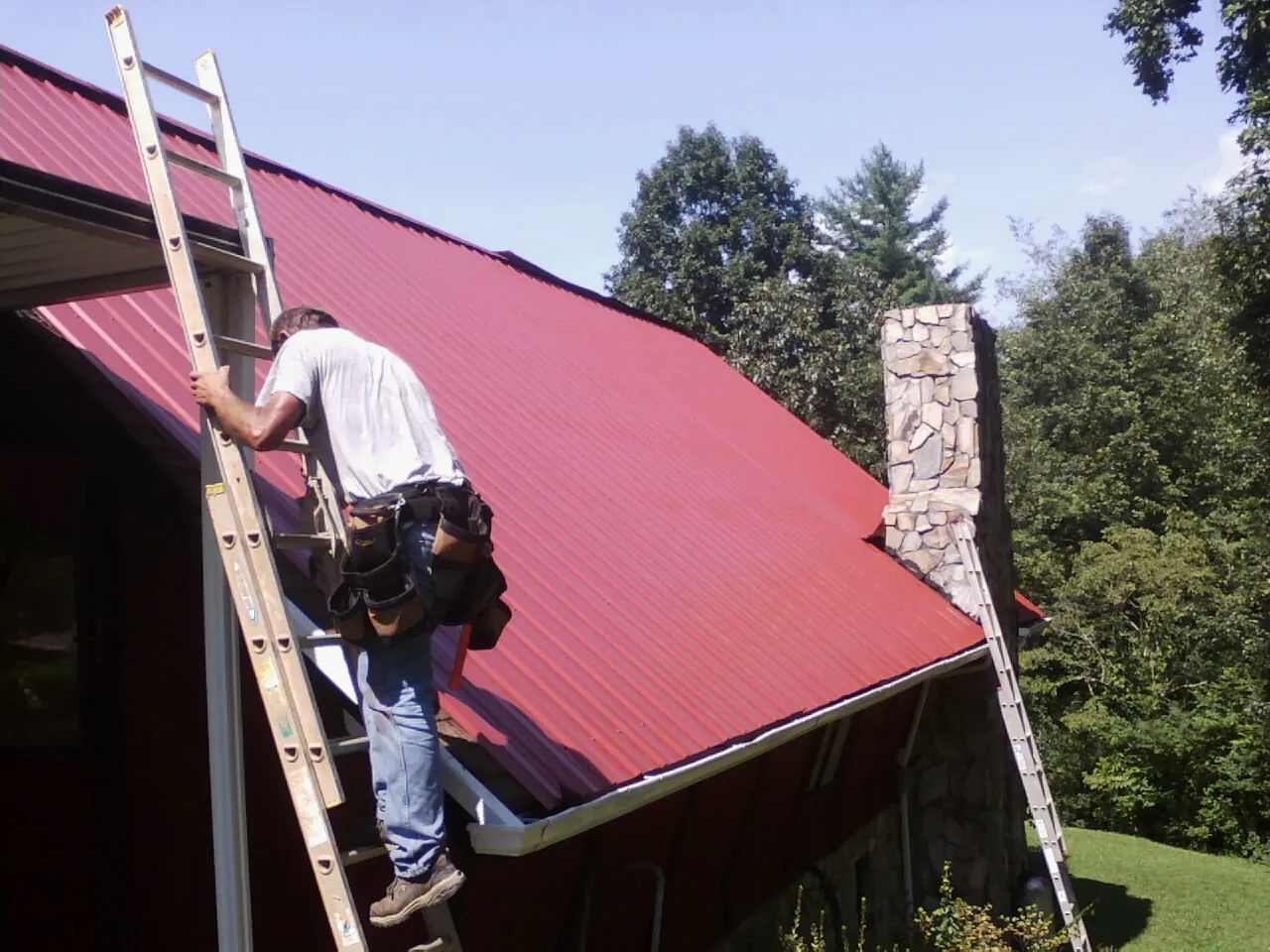 Roofer on a ladder beside a red metal roof and stone chimney — Soffit & Fascia Repair by Roofing Brothers in Pauls Valley